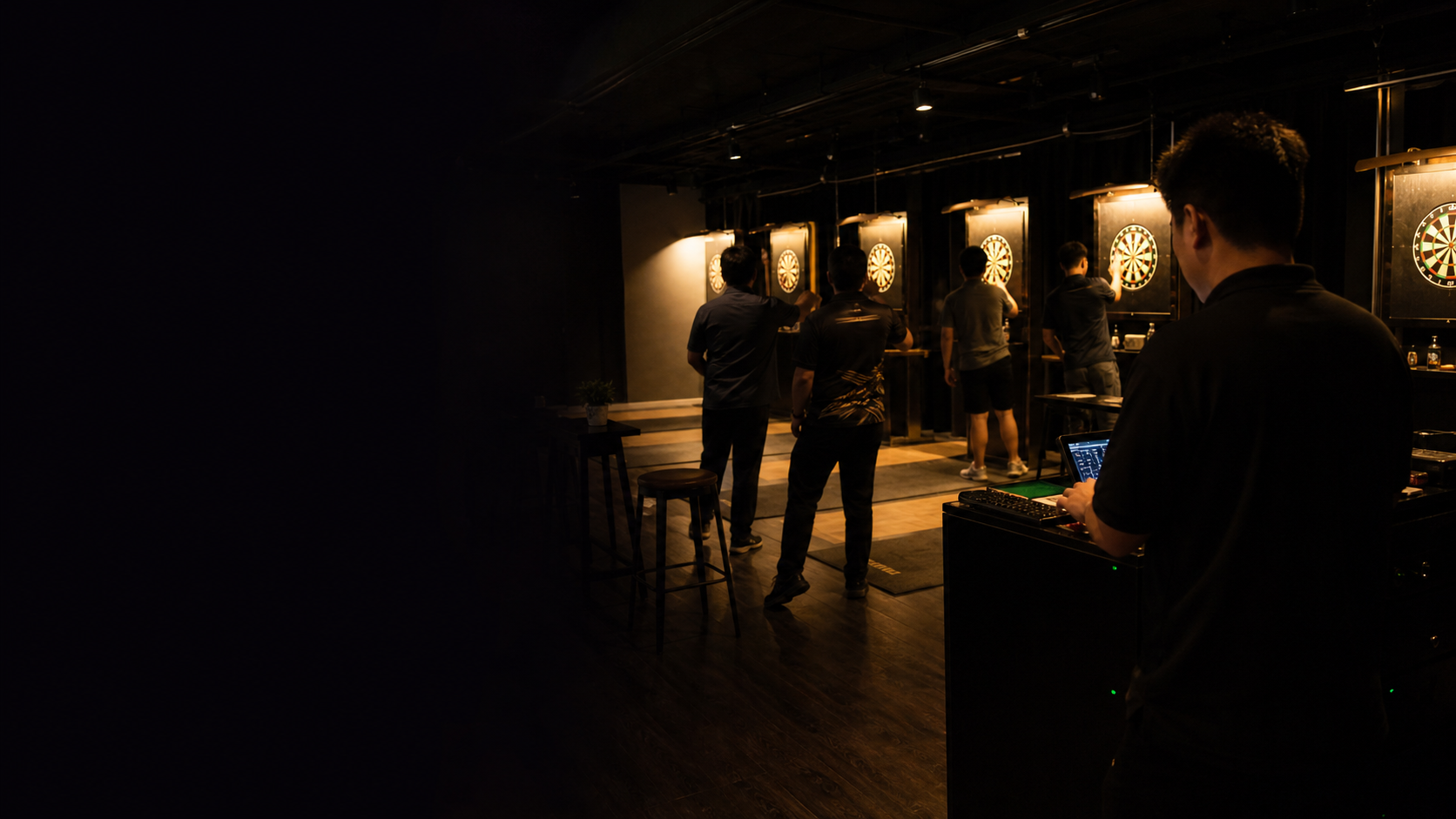 Darts venue at night with illuminated boards, players, and an operator at a control point.